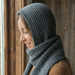 Könnte beinhalten: Ein graues Kapuzen- und Schal-Set aus Rippstrick. Die Kapuze bedeckt Kopf und Hals, und der Schal ist um den Hals gewickelt. Das Set ist aus einem weichen, strukturierten Garn gefertigt. Der Hintergrund ist eine Holzwand.