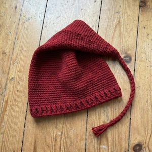 May include: A knitted red hat with a pointed crown and a long, plaited tie. The hat is made from textured yarn and rests on a wooden surface. The design of the hat is simple and classic.