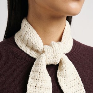 Puede incluir: Una bufanda de cuello tejida a ganchillo en color crema con un patrón de punto tejido texturizado. La bufanda está atada con un nudo sencillo en la parte delantera, sobre un suéter de punto de color burdeos oscuro con cuello redondo acanalado.