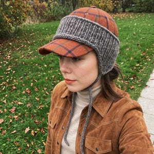 May include: A baseball cap featuring an orange and blue plaid pattern with a thick, grey, ribbed-knit wool band that covers the ears and forehead, complete with two long, grey, braided-knit ties hanging down from the sides.