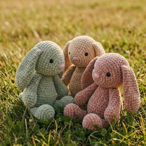 Peut inclure: Trois peluches de lapin crochetées, posées dans l'herbe. Les lapins sont de couleurs verte, beige et rose, avec des nez roses et des yeux noirs. Les peluches ont de longues oreilles tombantes et sont assises droites.