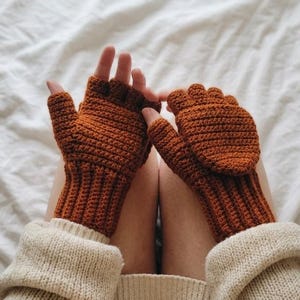 May include: A pair of burnt orange crochet fingerless gloves featuring a ribbed cuff and a fold-over mitten flap. The gloves are crafted with a textured stitch pattern and cover the hands while leaving the fingers exposed for dexterity.