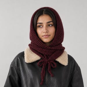 Op de afbeelding: Een diepbordeauxrode gebreide bivakmuts met een textuur van ribbelsteek. De capuchon heeft lange gebreide koorden die bij de nek zijn geknoopt en wordt gedragen over een zwart leren jack met een beige sherpa-kraag die het gezicht omlijst.
