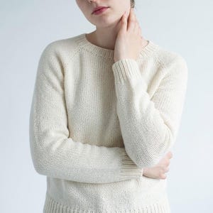 Könnte beinhalten: Ein langärmeliger, cremeweißer Strickpullover mit klassischem Rundhalsausschnitt und gerippten Details an Kragen, Ärmelbündchen und Saum. Das Kleidungsstück ist in einem gleichmäßigen glatten Rechtsstrick gefertigt, der eine klare Textur aufweist.