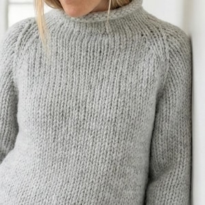 K&ouml;nnte beinhalten: Ein hellgrauer, grob gestrickter Pullover mit hohem, geripptem Kragen und gerippten B&uuml;ndchen. Der Pullover hat eine lockere Passform und ist aus einem weichen, strukturierten Garn gefertigt. Das Model tr&auml;gt dunkelblaue Jeans.