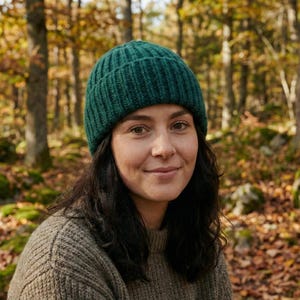 May include: A woman wearing a dark green knit beanie and a tan sweater, set against a blurred forest backdrop. The beanie is ribbed and fits snugly. The sweater has a textured appearance. The background features trees and fallen leaves.