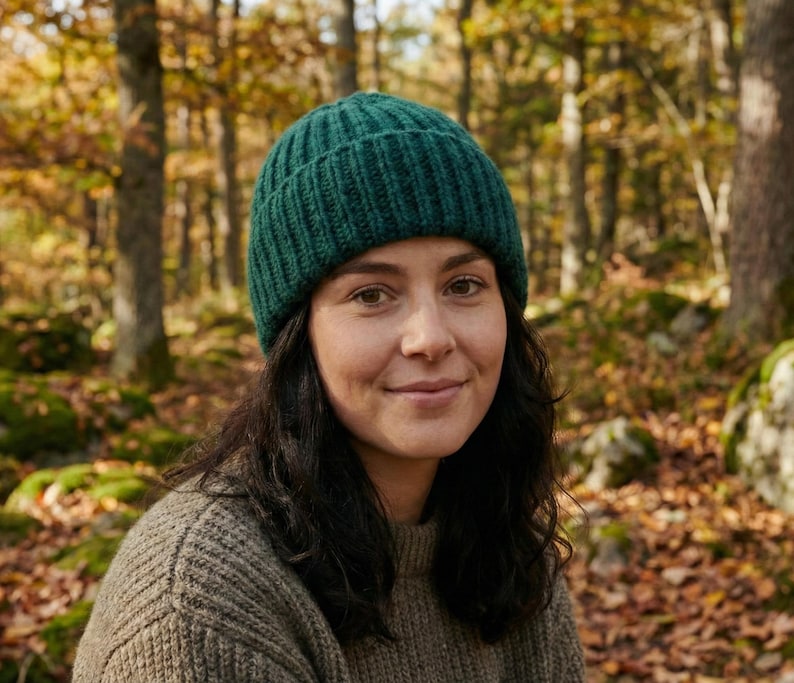 May include: A woman wearing a dark green knit beanie and a tan sweater, set against a blurred forest backdrop. The beanie is ribbed and fits snugly. The sweater has a textured appearance. The background features trees and fallen leaves.