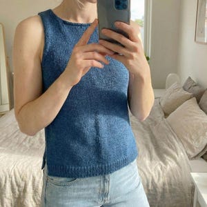 Könnte beinhalten: Ein ärmelloses, blaues Strickoberteil. Das Oberteil hat einen runden Ausschnitt und ein leicht strukturiertes Aussehen. Die Person auf dem Bild trägt hellblaue Jeans. Das Bild wurde in Innenräumen aufgenommen, mit einem Bett und einem Fenster im Hintergrund.
