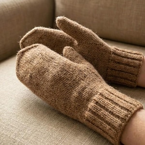 May include: A pair of brown knitted mittens. The mittens are made of a thick yarn and have a ribbed cuff. The mittens are shown on a person's hands, resting on a neutral-colored surface.