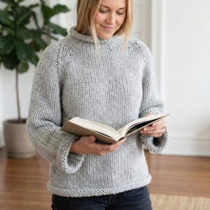 K&ouml;nnte beinhalten: Ein hellgrauer, grob gestrickter Pullover mit hohem Ausschnitt und langen &Auml;rmeln. Der Pullover ist aus dickem Garn gefertigt und hat ein strukturiertes Aussehen. Die Person h&auml;lt ein Buch.