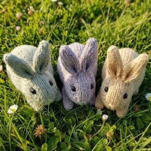 Op de afbeelding: Drie gebreide konijnen in groen, lila en beige, elk met zwarte knoopogen. De konijnen liggen op groen gras met kleine witte bloemen.