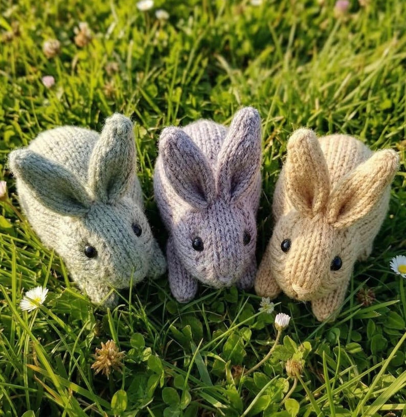 May include: Three knitted bunny rabbits in shades of green, purple, and tan, each with black button eyes. The bunnies are positioned on a bed of green grass with small white flowers.