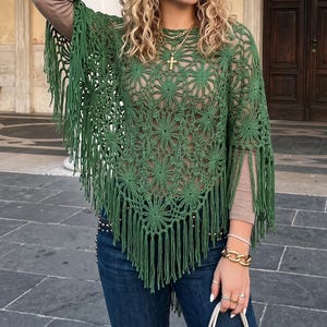 Puede incluir: Un poncho de ganchillo verde con flecos. El poncho presenta un patrón floral y se usa sobre una camiseta de manga larga y vaqueros azules. Incluye collares y pulseras doradas.