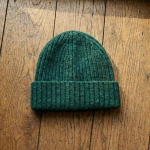 May include: A dark green ribbed knit beanie hat. The hat is made of a soft, textured material and features a folded brim. The beanie is a solid color and is suitable for cold weather.