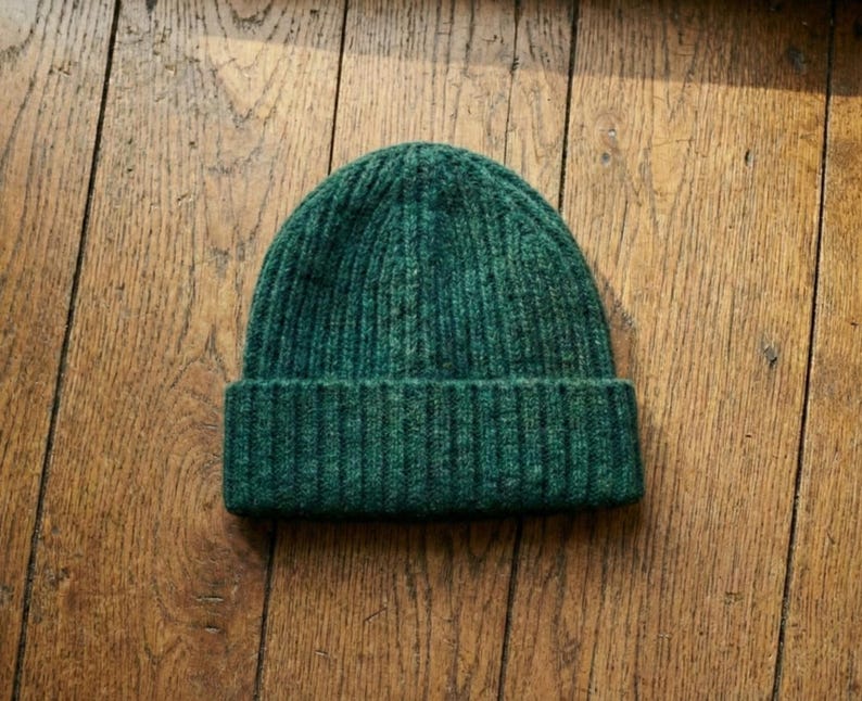 May include: A dark green ribbed knit beanie hat. The hat is made of a soft, textured material and features a folded brim. The beanie is a solid color and is suitable for cold weather.