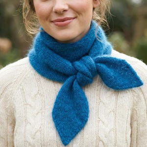 Può includere: Una sciarpa di lana blu brillante annodata sul davanti, con una finitura strutturata. La sciarpa è indossata sopra un maglione color crema a trecce. Le estremità della sciarpa sono a forma di foglia.