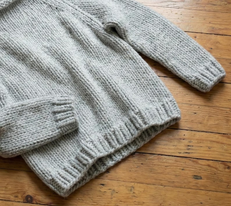 K&ouml;nnte beinhalten: Ein hellgrauer Strickpullover mit gerippten B&uuml;ndchen und Saum. Der Pullover hat lange &Auml;rmel und einen Rundhalsausschnitt. Der Strickstoff hat ein strukturiertes Aussehen. Der Pullover ist aus einem weichen, warmen Material gefertigt.