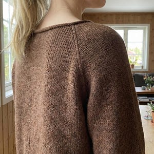 以下が含まれることがあります： クルーネックとラグランスリーブの茶色のニットセーター。セーターはテクスチャード加工の糸でできており、色は暖かくアースカラーです。画像はセーターの背面と肩を示しています。