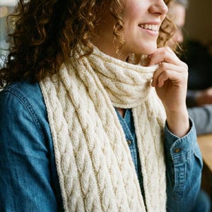May include: Cream-coloured cable knit scarf. The scarf is wrapped around the neck and shoulders. The scarf has a thick, textured appearance. The person is wearing a denim shirt.