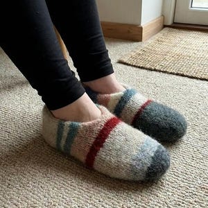 Puede incluir: Primer plano de un par de zapatillas de punto en una paleta de colores neutros. Las zapatillas presentan rayas horizontales rojas, azules y blancas. El tejido de punto suave y texturizado está diseñado para uso en interiores y comodidad.