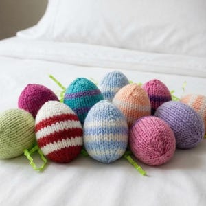 Puede incluir: Una colección de coloridos huevos de Pascua tejidos. Los huevos son de varios colores, incluyendo rosa, azul, verde y naranja, con diseños a rayas y lisos. Están dispuestos sobre una superficie blanca con hierba decorativa verde.