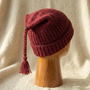 Puede incluir: Un gorro de punto burdeos con una larga borla trenzada y una peque&ntilde;a borla en el extremo. El gorro se exhibe sobre una forma de cabeza de madera sobre un fondo color crema. La textura del gorro es visible, mostrando el patr&oacute;n de punto.