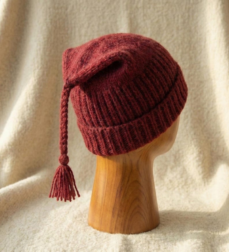 Puede incluir: Un gorro de punto burdeos con una larga borla trenzada y una peque&ntilde;a borla en el extremo. El gorro se exhibe sobre una forma de cabeza de madera sobre un fondo color crema. La textura del gorro es visible, mostrando el patr&oacute;n de punto.