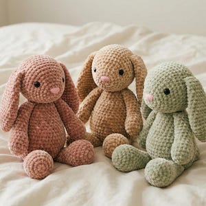 Könnte beinhalten: Drei handgefertigte Häkelhasen in Rosa-, Beige- und Grüntönen. Jeder Hase hat lange Schlappohren, einen runden Körper und eine kleine rosa Nase. Die Hasen sitzen auf einer weißen, strukturierten Oberfläche.