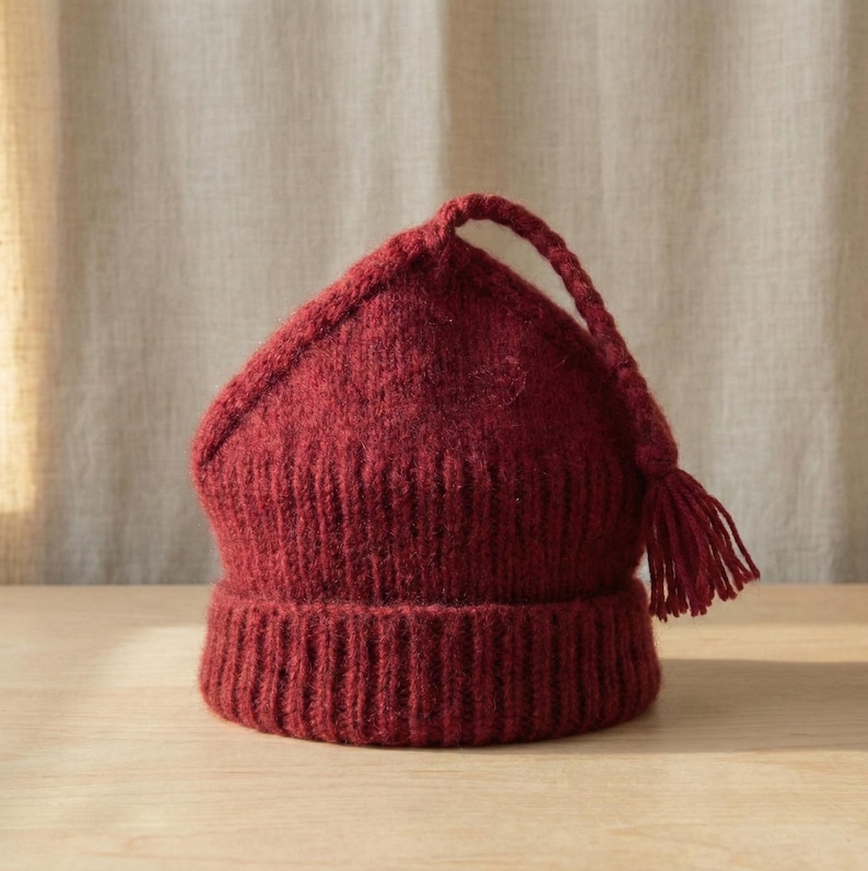 Puede incluir: Un gorro de punto burdeos con la parte superior puntiaguda y una borla. El gorro tiene un borde acanalado y un cord&oacute;n trenzado que se extiende desde la parte superior, terminando en una borla. El gorro est&aacute; hecho de un hilo suave y texturizado.