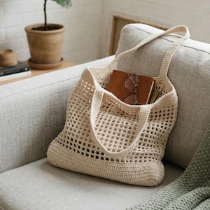 May include: A cream-colored, crocheted tote bag with a rectangular base and two handles. The bag is filled with a brown notebook, eyeglasses, and keys. The bag is sitting on a gray couch.