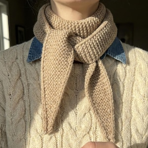 May include: A beige knitted scarf is tied around the neck, with the ends hanging down. The scarf is made of a thick, textured yarn. The person is wearing a cream-colored cable-knit sweater and a denim shirt underneath.