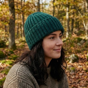 May include: A dark green ribbed knit beanie. The hat is made of a thick, warm material. The person is wearing a light brown sweater. The background is a forest with trees and fallen leaves.