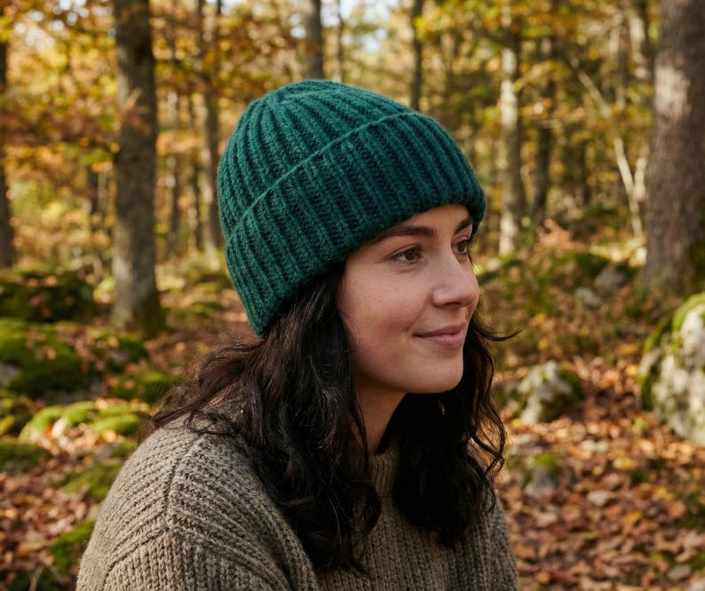 May include: A dark green ribbed knit beanie. The hat is made of a thick, warm material. The person is wearing a light brown sweater. The background is a forest with trees and fallen leaves.