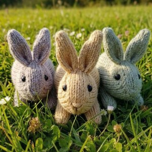 May include: Three knitted bunny rabbits in shades of gray, tan, and green sit in a bed of green grass. Each bunny has black eyes and long ears. The bunnies are handmade and appear to be soft and cuddly.