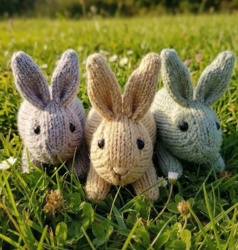 May include: Three knitted bunny rabbits in shades of gray, tan, and green sit in a bed of green grass. Each bunny has black eyes and long ears. The bunnies are handmade and appear to be soft and cuddly.