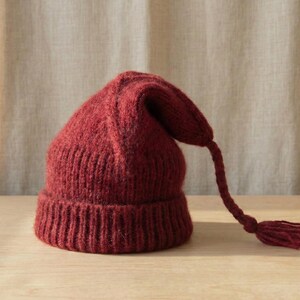 Puede incluir: Un gorro de punto burdeos con una larga borla trenzada. El gorro tiene un borde acanalado y una parte superior puntiaguda. La borla cuelga de la parte superior del gorro. El gorro est&aacute; hecho de un hilo suave y texturizado.