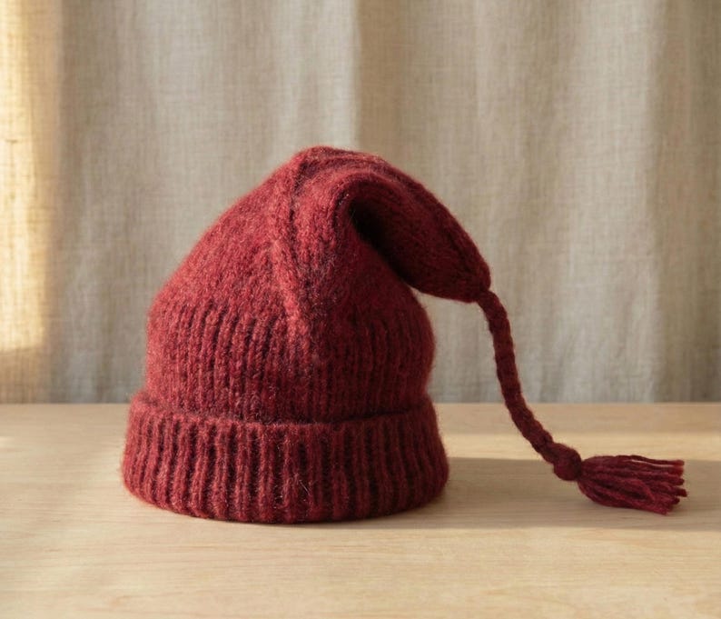 Puede incluir: Un gorro de punto burdeos con una larga borla trenzada. El gorro tiene un borde acanalado y una parte superior puntiaguda. La borla cuelga de la parte superior del gorro. El gorro est&aacute; hecho de un hilo suave y texturizado.