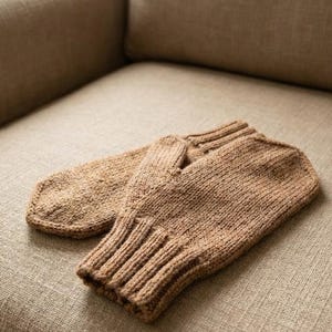 May include: A pair of knitted brown mittens. The mittens are made of a textured yarn and have a ribbed cuff. They are lying on a neutral-colored fabric surface.