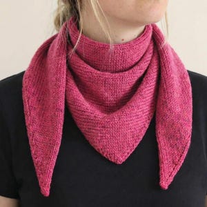 Peut inclure: Écharpe tricotée de couleur fuchsia vif. L'écharpe est de forme triangulaire, avec un motif de tricot texturé. Elle est drapée autour du cou, les extrémités pointant vers le bas. Le fond est noir uni.