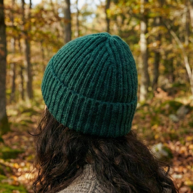 May include: A dark green ribbed knit beanie. The hat is made of a thick, warm material and is designed to keep the head warm. The beanie is shown from the back, with the wearer's dark hair and a neutral-colored sweater visible.