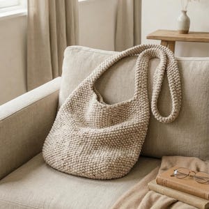 May include: A beige, knitted shoulder bag with a long strap rests on a beige sofa. The bag has a rounded shape and a textured surface. The background includes a window with beige curtains and a small wooden table.