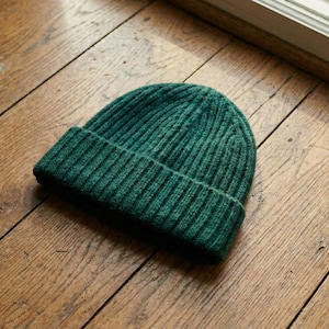 May include: A dark green ribbed knit beanie hat. The hat is made of a soft, textured material and has a folded brim. The beanie is a solid color and is sitting on a wooden surface.
