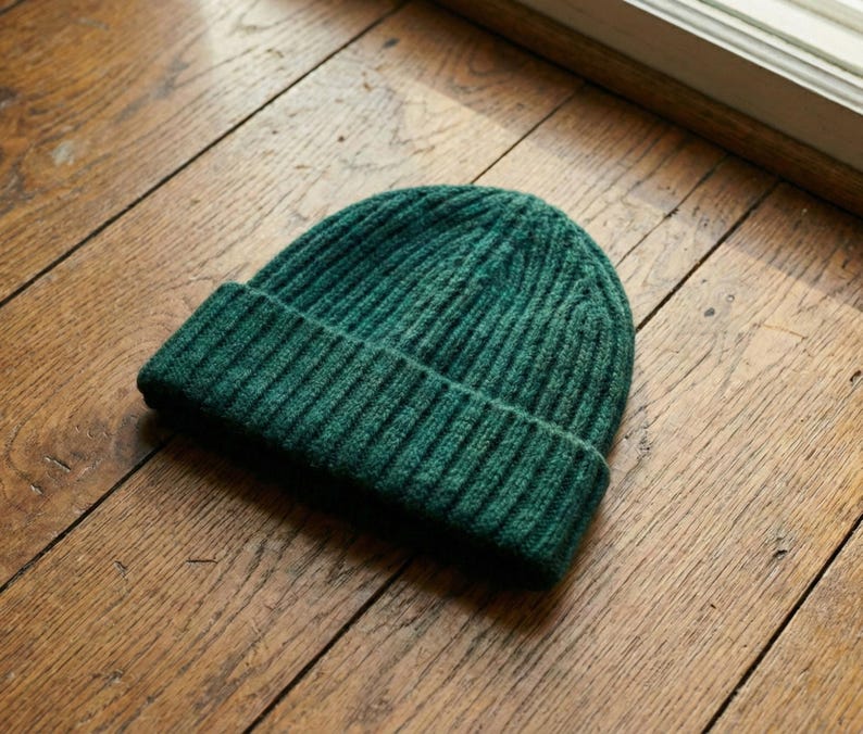 May include: A dark green ribbed knit beanie hat. The hat is made of a soft, textured material and has a folded brim. The beanie is a solid color and is sitting on a wooden surface.