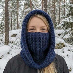 May include: A navy blue crocheted hood and face covering. The hood is up, and the face covering is pulled up to the nose. The background is a snowy forest.
