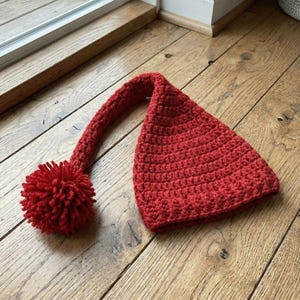 May include: A red crochet hat with a long, curved tail and a large pom-pom. The hat is made from textured yarn and is displayed on a wooden surface.