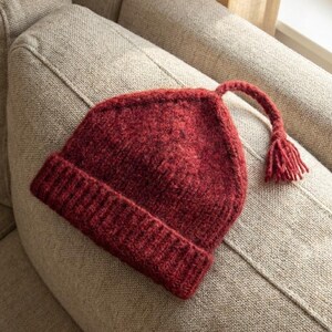 Puede incluir: Un gorro tejido de color burdeos con un borde doblado y una borla. El gorro est&aacute; hecho de un hilo texturizado y descansa sobre un sof&aacute; beige. El dise&ntilde;o del gorro es sencillo y cl&aacute;sico.