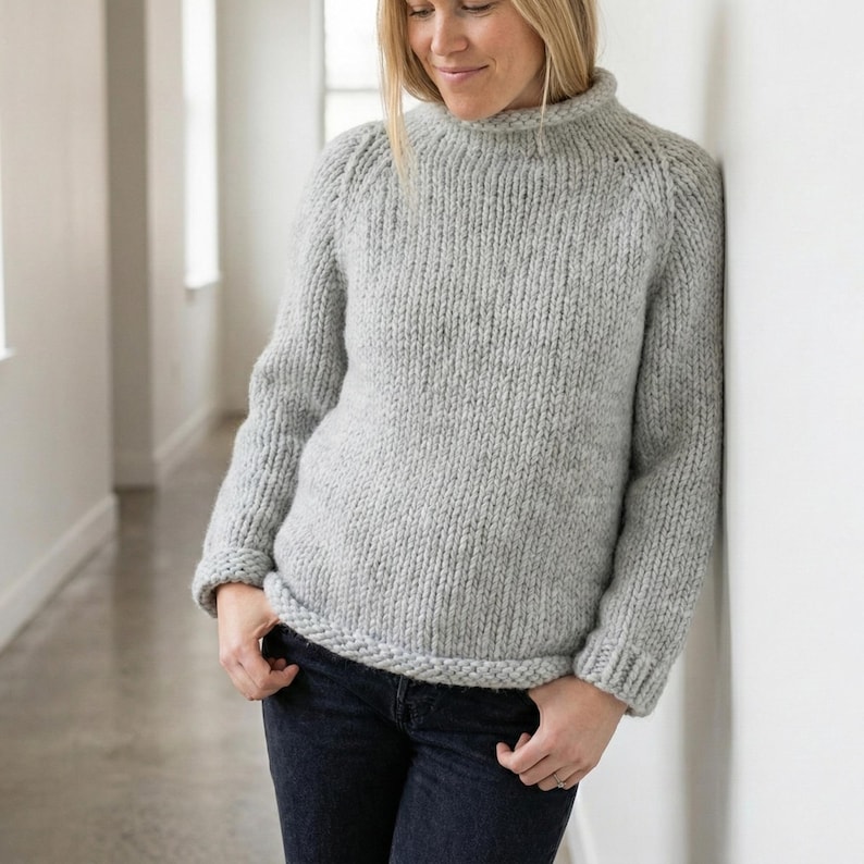 K&ouml;nnte beinhalten: Ein hellgrauer, grob gestrickter Pullover mit hohem, geripptem Kragen und gerippten B&uuml;ndchen. Der Pullover hat eine lockere Passform und ist aus einem weichen, strukturierten Garn gefertigt. Das Model tr&auml;gt dunkelblaue Jeans.