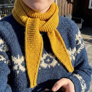 May include: A mustard yellow knitted scarf is draped over a blue sweater with a white snowflake pattern. The scarf is knotted at the neck and hangs down. The sweater has a crew neck and long sleeves.