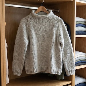 K&ouml;nnte beinhalten: Ein hellgrauer, grob gestrickter Pullover mit hohem, geripptem Ausschnitt. Der Pullover h&auml;ngt an einem Holzb&uuml;gel in einem Holzschrank. Der Schrank hat Regale mit gefalteter Kleidung.