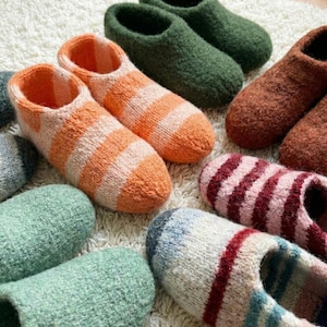 Pode incluir: Uma coleção de pantufas de malha feitas à mão em várias cores e padrões. As pantufas incluem designs listrados em laranja e branco, verde, marrom e rosa e vermelho. O calçado é disposto sobre uma superfície branca texturizada.